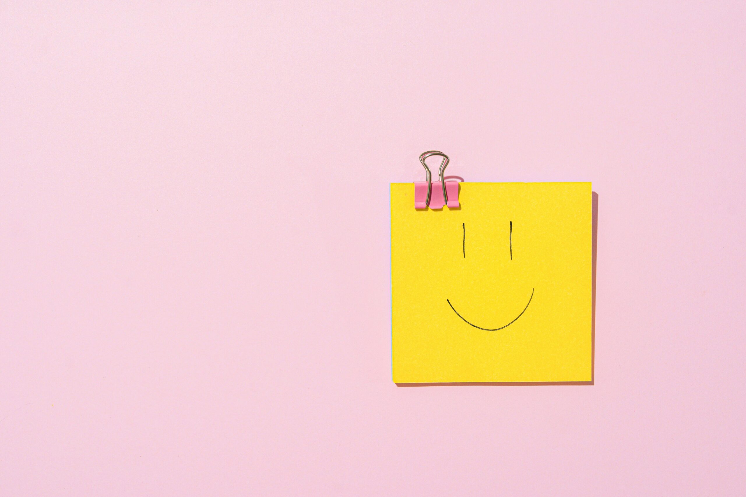 Yellow sticky note with a smiley face clipped to a pink surface, conveying happiness and simplicity.